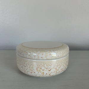 May include: A round, lidded ceramic container with a cream-colored base and lid. The container features a speckled, brown pattern across its surface. The lid is flat and the container is sitting on a white surface.