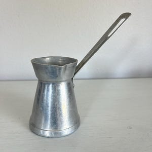 May include: A silver-colored metal coffee pot with a wide, flared rim, a small pouring spout, and a long, flat, angled handle. The vessel has a tapered cylindrical body with a smooth, metallic finish and a visible rivet connecting the handle.