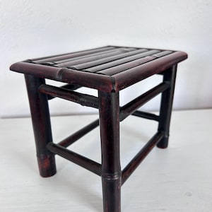 May include: A small, dark red bamboo stool with a rectangular seat. The seat is made of several bamboo slats. The legs are sturdy and angled, connected by horizontal and diagonal supports. The stool is a traditional Asian-inspired piece.