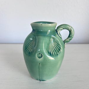 May include: A small, turquoise ceramic pitcher with a handle. The pitcher features seashell designs on the front and a floral design. The handle has a twisted rope design. The pitcher is on a white surface.