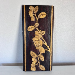May include: A rectangular wooden wall art piece with a dark burgundy background and a gold floral design. The floral design is carved and features flowers and leaves. The edges of the piece are also trimmed in gold.