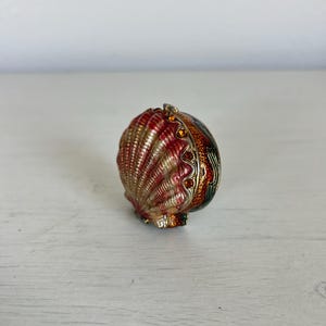 May include: A small, hinged trinket box shaped like a seashell, featuring a metallic red and gold finish with textured ridges. The edge is decorated with a row of small, amber-colored crystals along the seam of the shell casing.