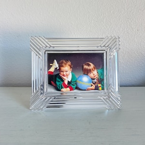 May include: A rectangular picture frame made of clear, molded glass with a ribbed, stepped design along the edges. The frame contains a photograph of two children, one resting their chin on their hand and the other holding a blue ball.