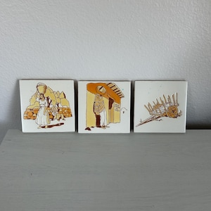May include: Three square ceramic tiles with illustrations in shades of brown and yellow. The tiles depict scenes of people and objects, possibly from an ancient civilization. The artwork is outlined in black on a white background.