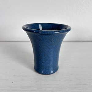 May include: A small, blue ceramic vase with a speckled texture. The vase has a flared top and a cylindrical body. The color is a deep, rich blue with darker speckles throughout. The vase is likely handmade.