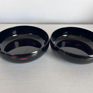 May include: Two black, glossy bowls with a rounded, asymmetrical design. The bowls have a deep, dark color with subtle red accents visible on the rim. The bowls are placed side by side.