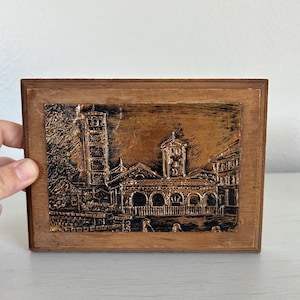 May include: A rectangular wooden plaque featuring a detailed relief of a building with a tower and arched windows. The relief is in shades of brown and copper, set within a dark brown wooden frame. The artwork depicts a historical architectural scene.