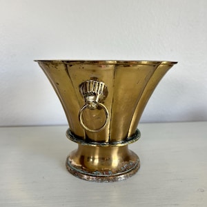 May include: A polished brass vessel with a flared, fluted rim and a pedestal base. The exterior features a decorative lion head motif on each side, integrated with a circular ring handle. The metal shows signs of natural patina and oxidation.