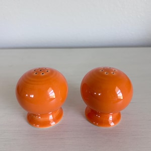 May include: Two orange ceramic salt and pepper shakers. Each shaker is spherical with a small, circular base and a series of holes at the top. The shakers have a glossy finish and are displayed on a light-colored surface.