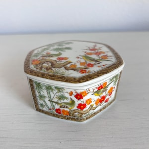 May include: A hexagonal porcelain trinket box with a lid. The box features a detailed design of birds, flowers, and bamboo. The lid and sides are decorated with a brown border. The color palette includes white, green, orange, and brown.