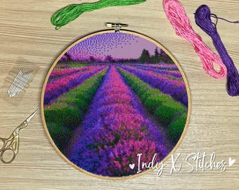 May include: A close-up of a cross-stitch embroidery hoop with a colorful image of a lavender field. The field is a mix of purple, pink, and green hues, with a path leading through the center. The hoop is made of wood and has a natural finish.