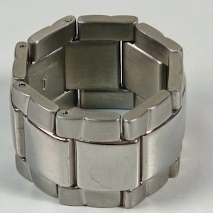 May include: A silver-toned metal bracelet with a geometric design. The bracelet is composed of rectangular and curved segments, connected to form a flexible band. The surface has a brushed finish, and the overall style is modern and minimalist.