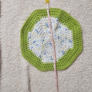 May include: A green and white crocheted coaster with a circular design. The coaster is approximately 15 cm in diameter.