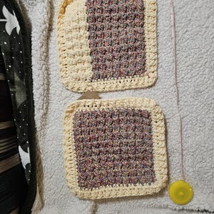 May include: Two crocheted dishcloths, one with a purple, pink, and yellow yarn and the other with a white yarn. Both dishcloths have a cream-colored border.