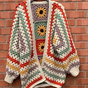 May include: A crochet cardigan constructed from multi-coloured granny squares featuring sunflower motifs in yellow and brown, accented by stripes of cream, sage green, burnt orange, and muted purple, finished with cream-coloured ribbed cuffs.