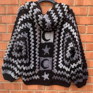 May include: A hooded crochet jumper featuring a black and grey granny square pattern. The back panel displays a vertical arrangement of square motifs with alternating crescent moon and star designs in shades of black and grey yarn.