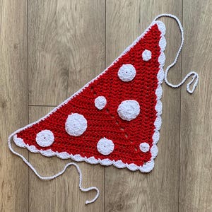 May include: A red and white crochet bandana with a mushroom design. The bandana is triangular, with a red base and white polka dots. The edges are trimmed with a white scalloped border and white ties.