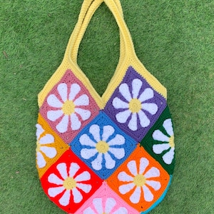 May include: A colourful, handmade crochet tote bag with a daisy pattern. The bag has a yellow handle and trim, and is made up of squares in various colours, including pink, orange, blue, and green, each with a white daisy. The bag is made of yarn.