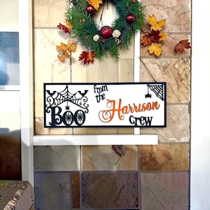 May include: A rectangular white sign with black and orange lettering and cutouts. The left side features the word Boo in black with spiderwebs and bats. The right side displays the text from the Harrison Crew in black and orange script.