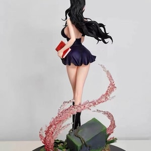 May include: A black haired anime figurine wearing a black dress and holding a red book. The figurine is standing on a base with green plants and a gray rock. The figurine is surrounded by pink and white translucent swirls.