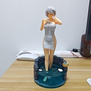 May include: A white figurine of a woman standing in a small pool of water. She is wearing a white towel and has her hands on her face. The pool is surrounded by a stone wall.