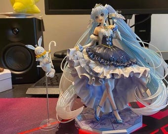 Puede incluir: Figura detallada de un personaje con largo cabello azul claro y un vestido blanco y azul marino. La figura está posada sobre una base hexagonal. También hay una pequeña figura de dos conejos blancos con detalles amarillos.
