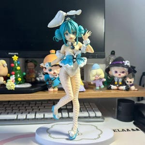 May include: A blue-haired anime figurine wearing a white bunny costume with blue accents. The figurine is standing on a white base with a gold trim. The figurine is posed with one hand raised and the other on her hip.