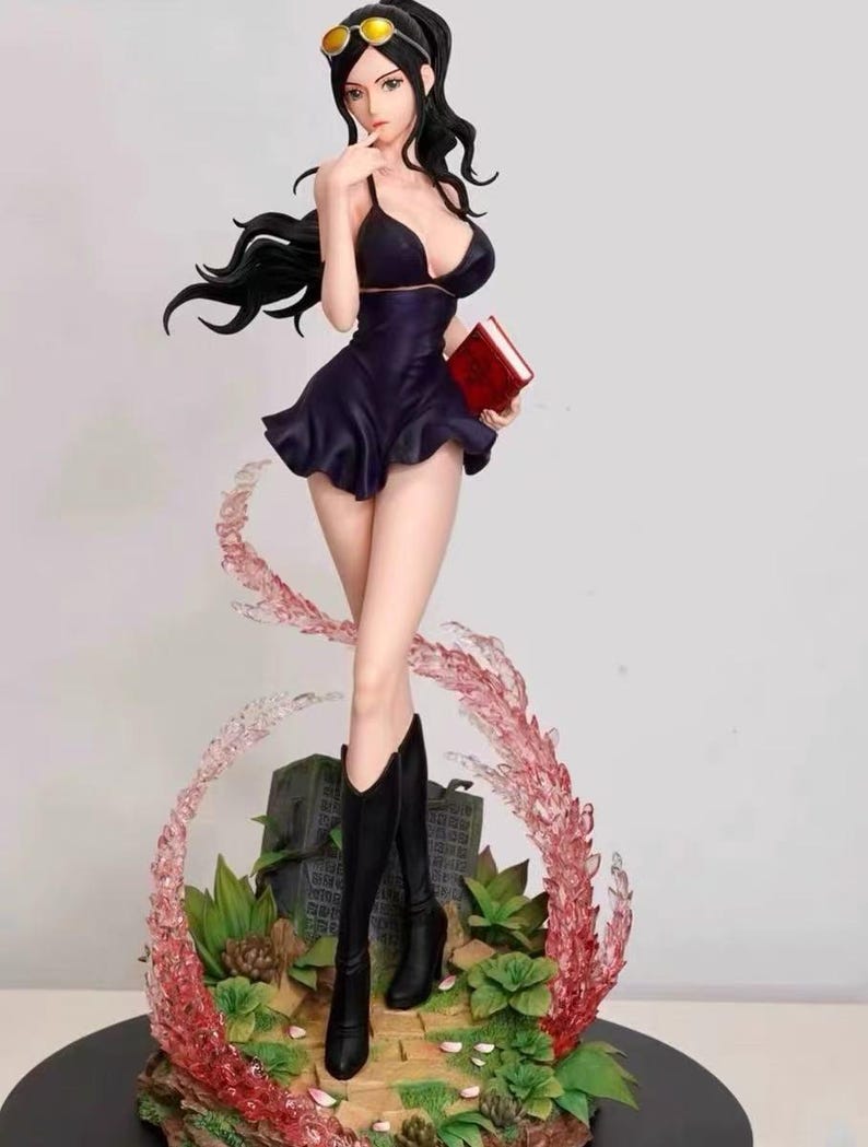 May include: A black and purple anime figurine of a woman with long black hair, wearing a black dress, sunglasses, and black boots. She is holding a red book and standing on a base with green plants and pink and white swirls.