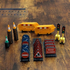 May include: A miniature construction set featuring yellow plastic barriers, orange and white traffic cones, traffic lights, and various road signs including red and white signs that read ROAD WORKS and ROAD CLOSED, with a 30 cm ruler for scale.