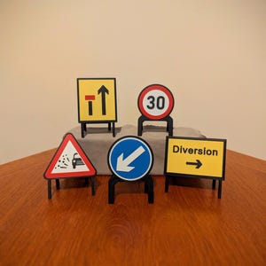 Può includere: Una collezione di segnali stradali in miniatura, tra cui un segnale di limite di velocità e un segnale di deviazione. I segnali sono neri, bianchi, rossi e gialli e sono esposti su una superficie di legno.