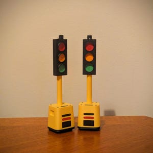 Può includere: Due semafori in miniatura. Ognuno ha una testa di segnale nera con luci rosse, gialle e verdi. Le teste dei segnali sono montate su pali gialli e poggiano su basi gialle con strisce nere e rosse. Su una superficie di legno.