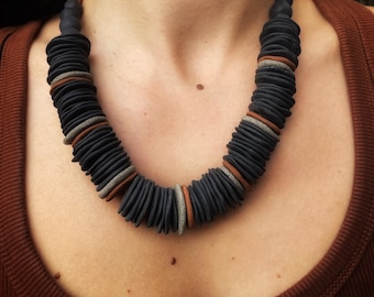 May include: A necklace featuring numerous small, flat, circular dark charcoal grey beads strung together. Interspersed among the dark beads are occasional thin, flat discs in terracotta orange and light grey, creating a repeating pattern.