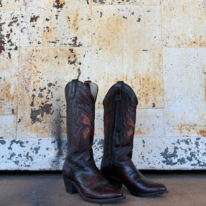 May include: A pair of dark brown leather cowboy boots featuring a pointed toe, a stacked heel, and decorative stitching in a reddish-brown hue on the tall shaft. The boots have a classic pull-on design with side pull tabs at the top opening.