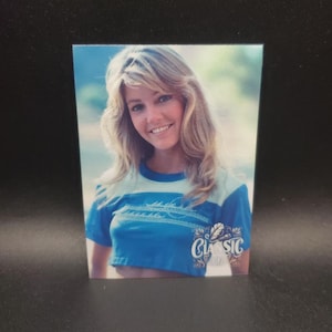 Puede incluir: Fotografía vintage de una persona sonriente con cabello rubio. La persona lleva un top corto azul con rayas blancas y la palabra "CLASSIC" en una fuente decorativa. El fondo está borroso.