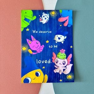 May include: A rectangular art print with a blue background featuring cartoon characters and yellow stars. The text "We deserve to be loved" is written in white. The characters are in various colors, including pink, green, and yellow.