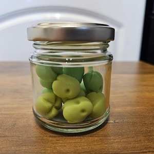 以下が含まれることがあります： 銀色の蓋が付いたガラス瓶に緑色のキャンディーが入っています。キャンディーは小さく丸い果物の形をしています。