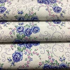 Op de afbeelding: Close-up van gevouwen stof met een bloemenpatroon. De stof heeft grote blauwe rozen, kleine roze bloemen en groene bladeren op een witte achtergrond. Het ontwerp bevat gestippelde wervelaccenten, wat een vintage, romantische uitstraling creëert.