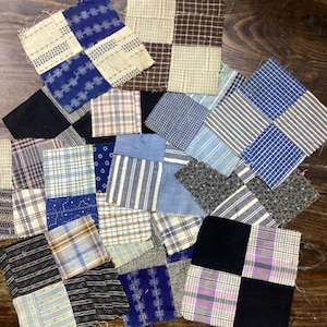Può includere: Una collezione di quadrati di tessuto assortiti in vari modelli e colori, tra cui blu, bianco, nero e a quadri. I quadrati sono disposti su una superficie di legno scuro, mostrando un design patchwork.