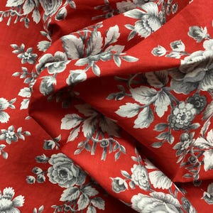 Puede incluir: Primer plano de tela roja con estampado floral. El diseño presenta grandes rosas grises y blancas y otras flores con hojas verdes. La tela está doblada, mostrando la textura y profundidad del material.