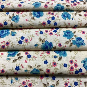 Peut inclure: Gros plan sur un tissu plié avec un motif floral. Le motif présente de grandes roses bleues, de petites fleurs roses et bleues, et des feuilles vertes sur un fond crème. Le tissu est plié en couches horizontales.