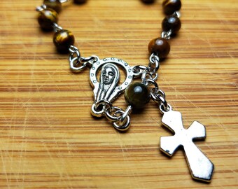 May include: A brown and gold tiger's eye beaded rosary with a silver charm featuring a depiction of the Virgin Mary and a silver cross.