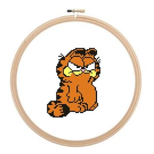 May include: Cross-stitch embroidery of Garfield the cat in an embroidery hoop. The orange cat with black stripes has an angry expression. The wooden hoop is round and has a metal clasp at the top.