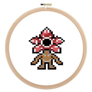 Könnte beinhalten: Ein Kreuzstich-Stickrahmen mit einem pixeligen Demogorgon aus der beliebten Science-Fiction-Serie. Der Demogorgon ist in Rot-, Schwarz- und Brauntönen vor einem weißen Hintergrund dargestellt. Der Stickrahmen ist aus hellem Holz gefertigt.
