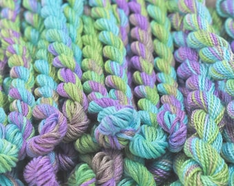 Könnte beinhalten: Nahaufnahme einer Sammlung von Stickgarn in Grün-, Blau- und Violetttönen. Das Garn ist in einem zufälligen Muster angeordnet, wodurch ein strukturiertes und farbenfrohes Display entsteht.