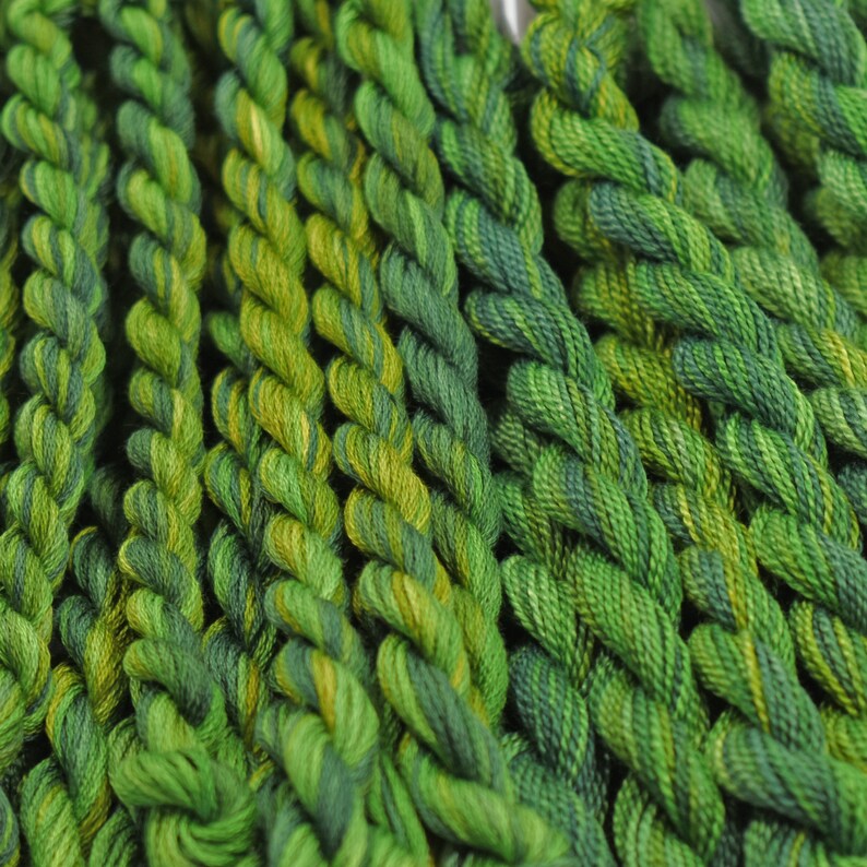 May include: A collection of green and yellow embroidery floss spools. The floss is arranged in rows, with each row featuring a different shade of green.