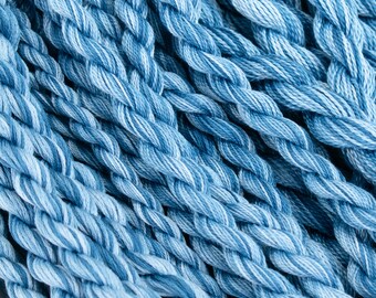 Puede incluir: Un manojo de hilo de bordar de algodón trenzado en azul y blanco. Las hebras presentan un patrón de color jaspeado con tonos alternos de azul claro y medio, creando una apariencia texturizada similar a una cuerda en toda la superficie.