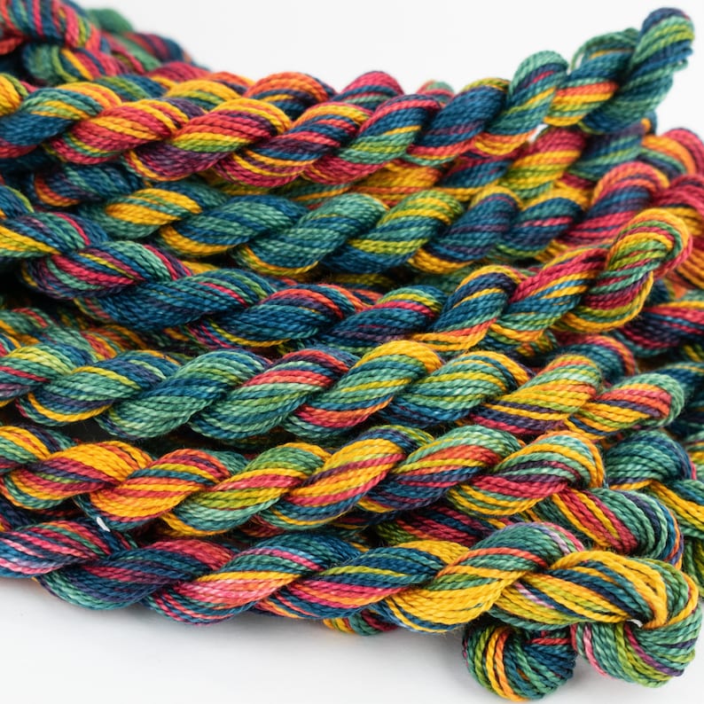 May include: A close-up of a pile of colorful embroidery floss. The floss is a mix of red, orange, yellow, green, blue, and purple. The floss is arranged in a braided pattern.