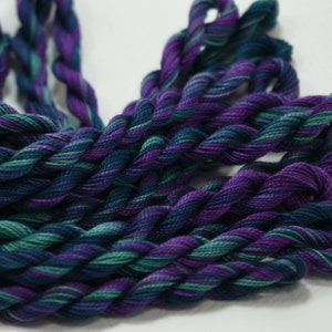 May include: Close-up of several skeins of embroidery floss in shades of purple, teal, and dark blue. The threads are twisted together, creating a textured appearance. The floss is likely used for cross-stitch, embroidery, or other needlecraft projects.