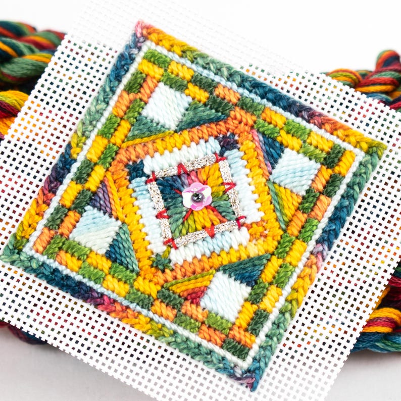 May include: A colorful cross-stitch pattern with a geometric design. The pattern is worked on Aida cloth and features a variety of colors, including yellow, orange, green, blue, and red. A small, pink button is centered in the design.