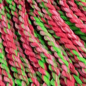 May include: Close-up of a bundle of embroidery floss in shades of pink, green, and red. The floss is arranged in a criss-cross pattern, creating a textured and colourful background.